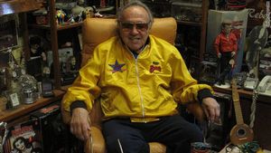 An old man in a bright yellow jacket and large sunglasses sits comfortably in a chair. In the background, numerous souvenirs from his old days are present.