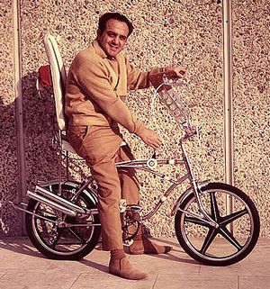 A 1967 photograph showing George Barris in a custom-made bicycle by the wall smiling. The picture looks funny, because Barris is already past his thirtees while the bike looks like it belongs to a teenager.