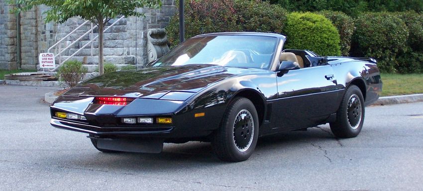 KITT seen from profile, with its middle red light on and other lights off. Some buildings and nature in the background.