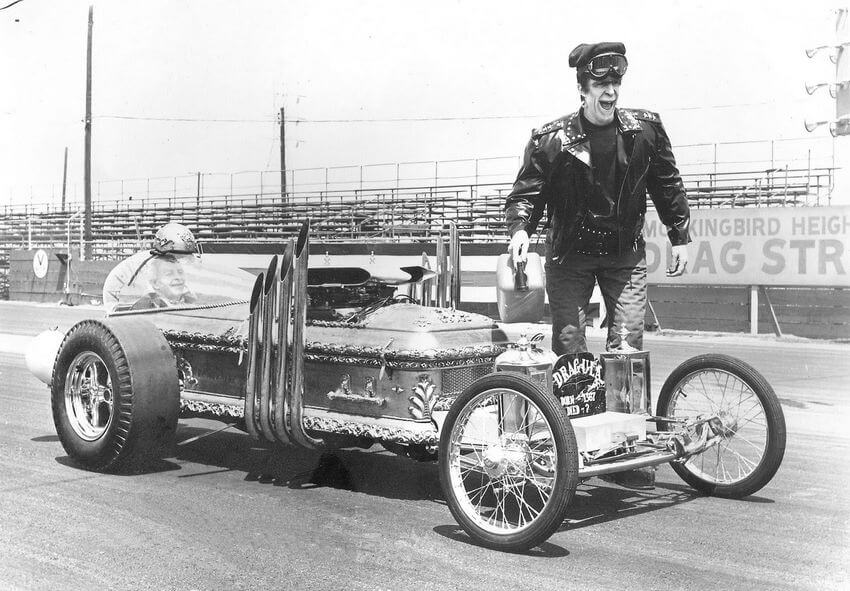 Black and white picture showing two main stars of the show having a laugh, with Grandpa inside his car and Herman standing by the car, touching it.