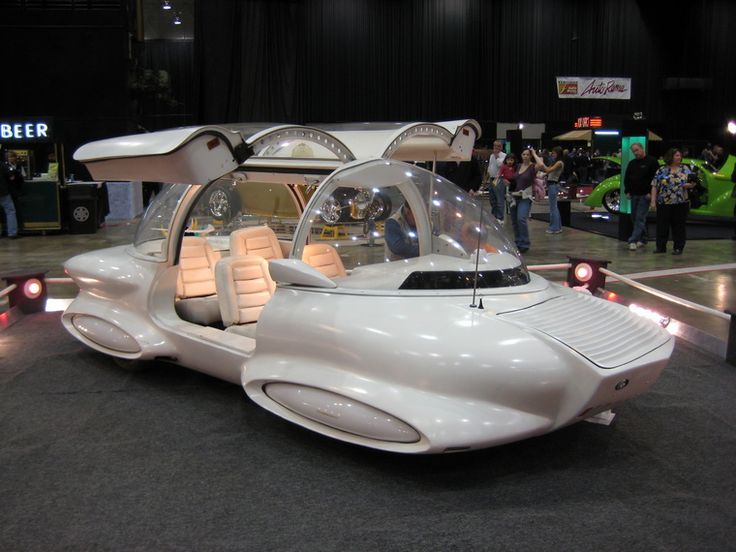 White and bubbly car with doors opened vertically and lights on inside, illuminating bright passenger seats.