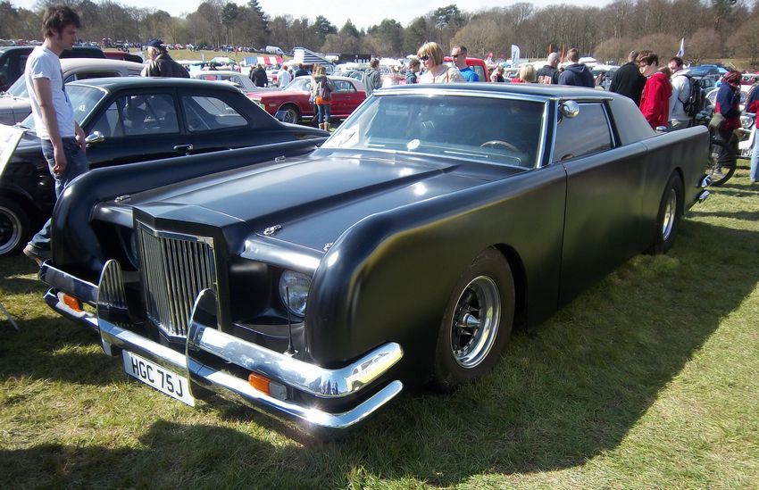 Big and bulky black car on some open air car show, with lots of people looking around and appreciating various vehicles presented.