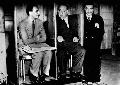 Alfred Hitchcock with his writer and Peter Llore standing in what looks like a stripped confessional.