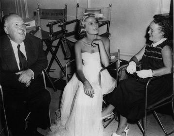 Alma Reville, Grace Kelly and Hitchcock looking at someone else attentively. Grace Kelly is in a beautiful white wedding dress.