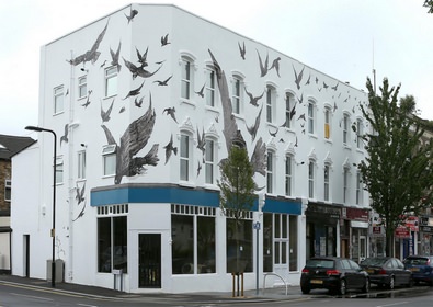 Many black birds flying cover the paint of two sides of a building positioned on the corner.