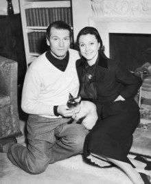 Laurence Olivier and Vivien Leigh sit on the floor and petting their cat New Boy who sit at their lap and seems visibly scared of something.