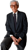 Lew Wasserman sitting on a desk, with his iconic glasses on, half-smiling.